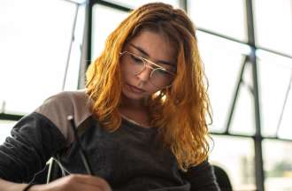 [Featured image] A young woman with orange hair and glasses concentrates as she writes. 