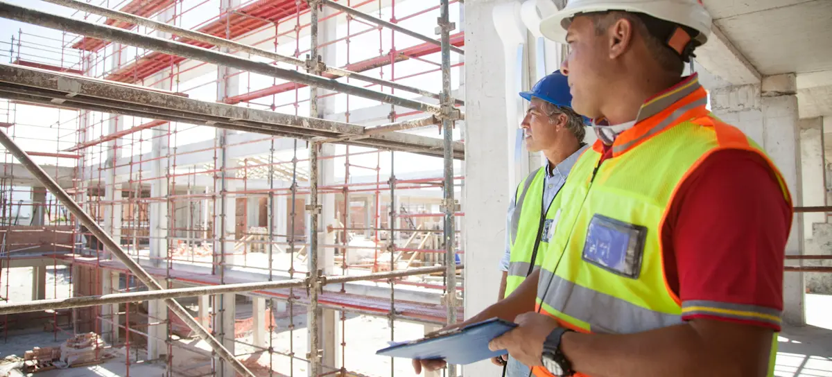 [Featured image] Two construction workers gaze out at a construction site; one holds a tablet with information protected by strategies enhancing cybersecurity in the construction industry.
