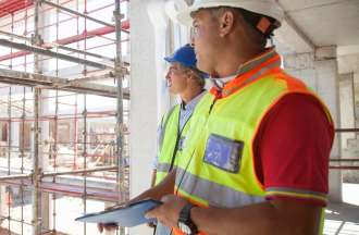 [Featured image] Two construction workers gaze out at a construction site; one holds a tablet with information protected by strategies enhancing cybersecurity in the construction industry.
