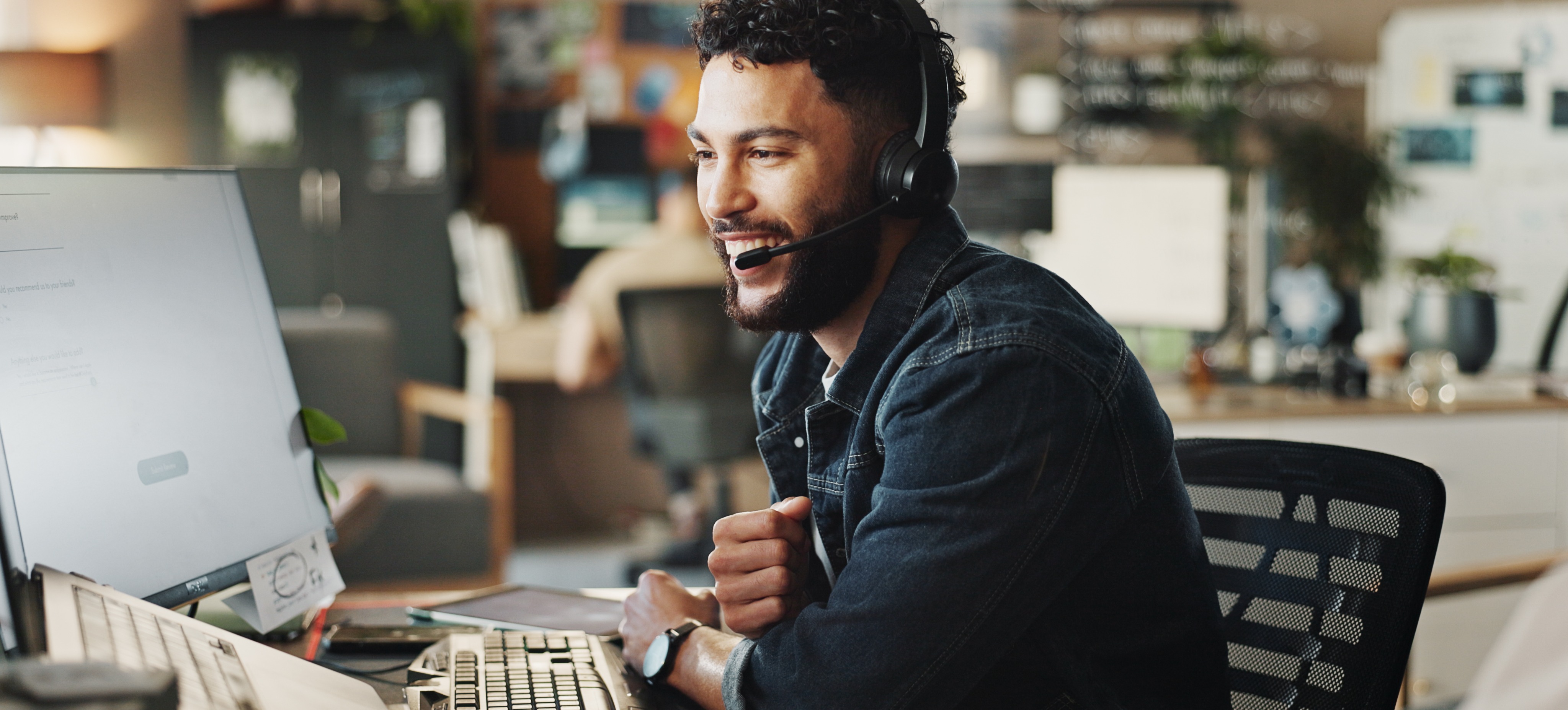 [Featured Image] A smiling technical support analyst speaks through a headset and looks at his computer monitor and equipment to troubleshoot a client's IT issues.
