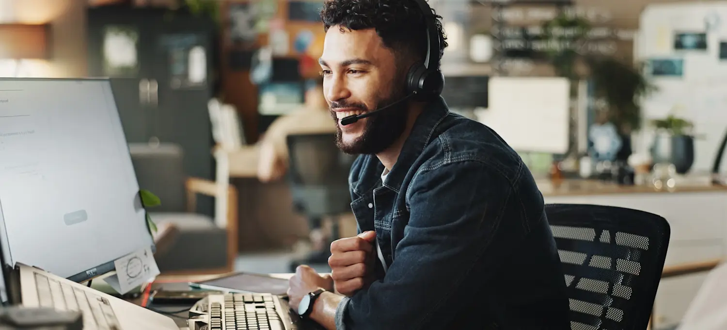 [Featured Image] A smiling technical support analyst speaks through a headset and looks at their computer monitor and equipment to troubleshoot a client's IT issues.
