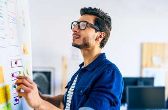 [Featured image] A UX designer wearing eyeglasses and a blue shirt draws a wireframe on a whiteboard.