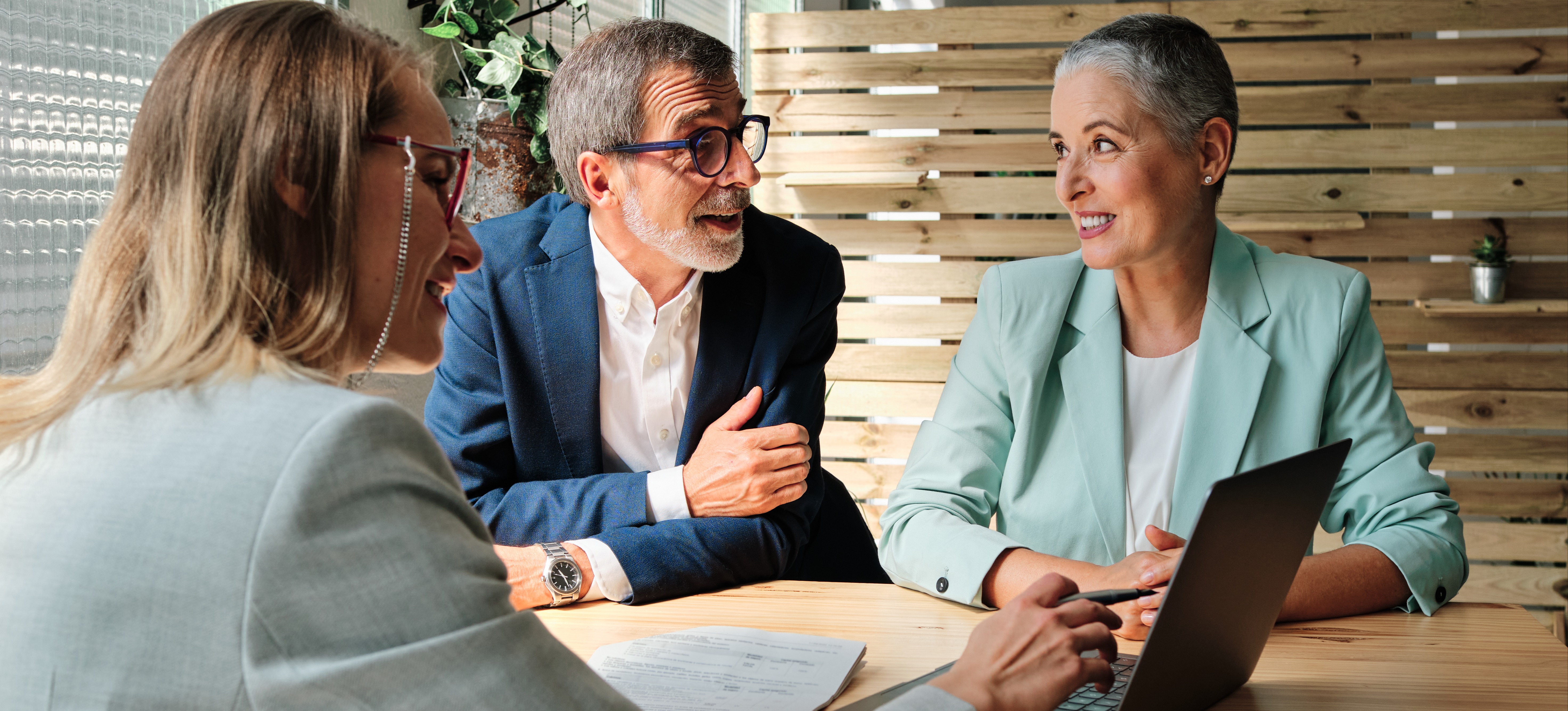 [Featured Image] A personal finance advisor talks to two clients at a table, showing them their financial data on a laptop.