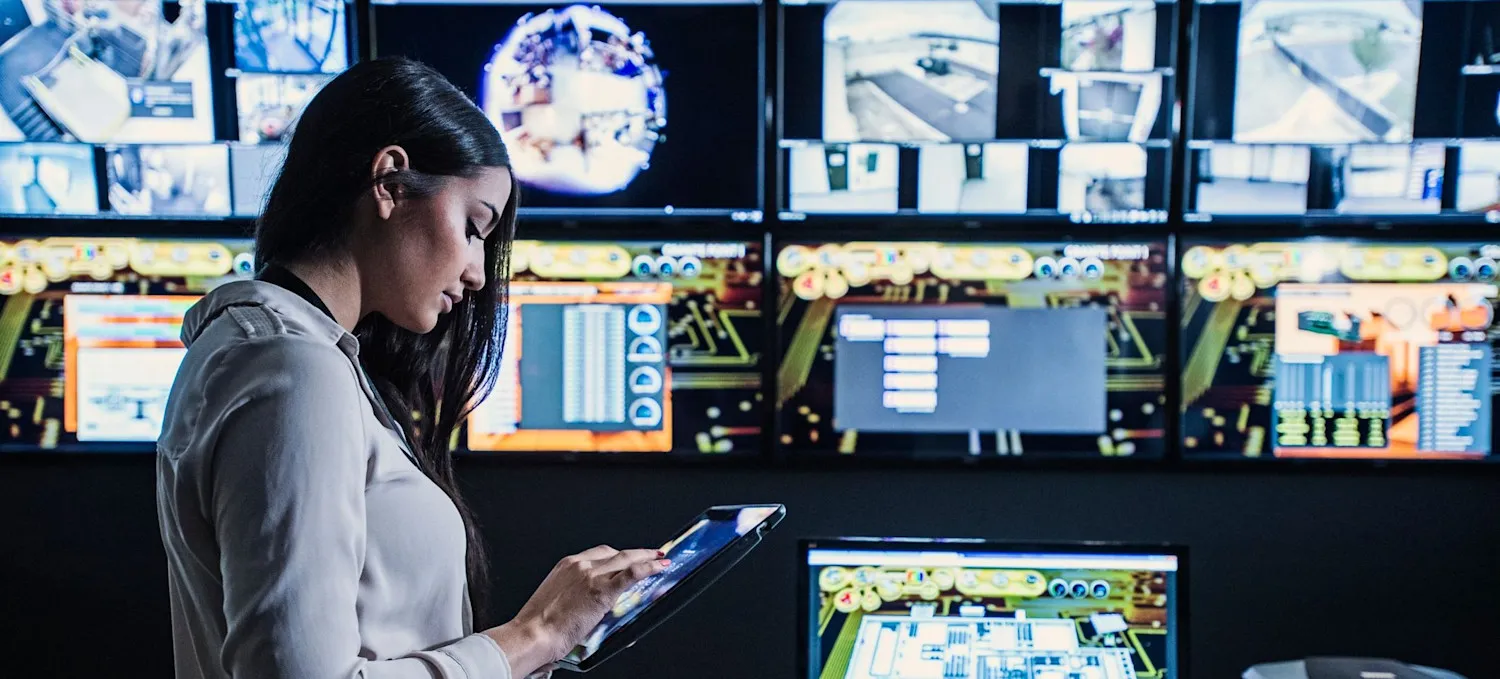 [Featured Image] A person stands in a darkened room lined with video monitors and reviews information on a tablet.