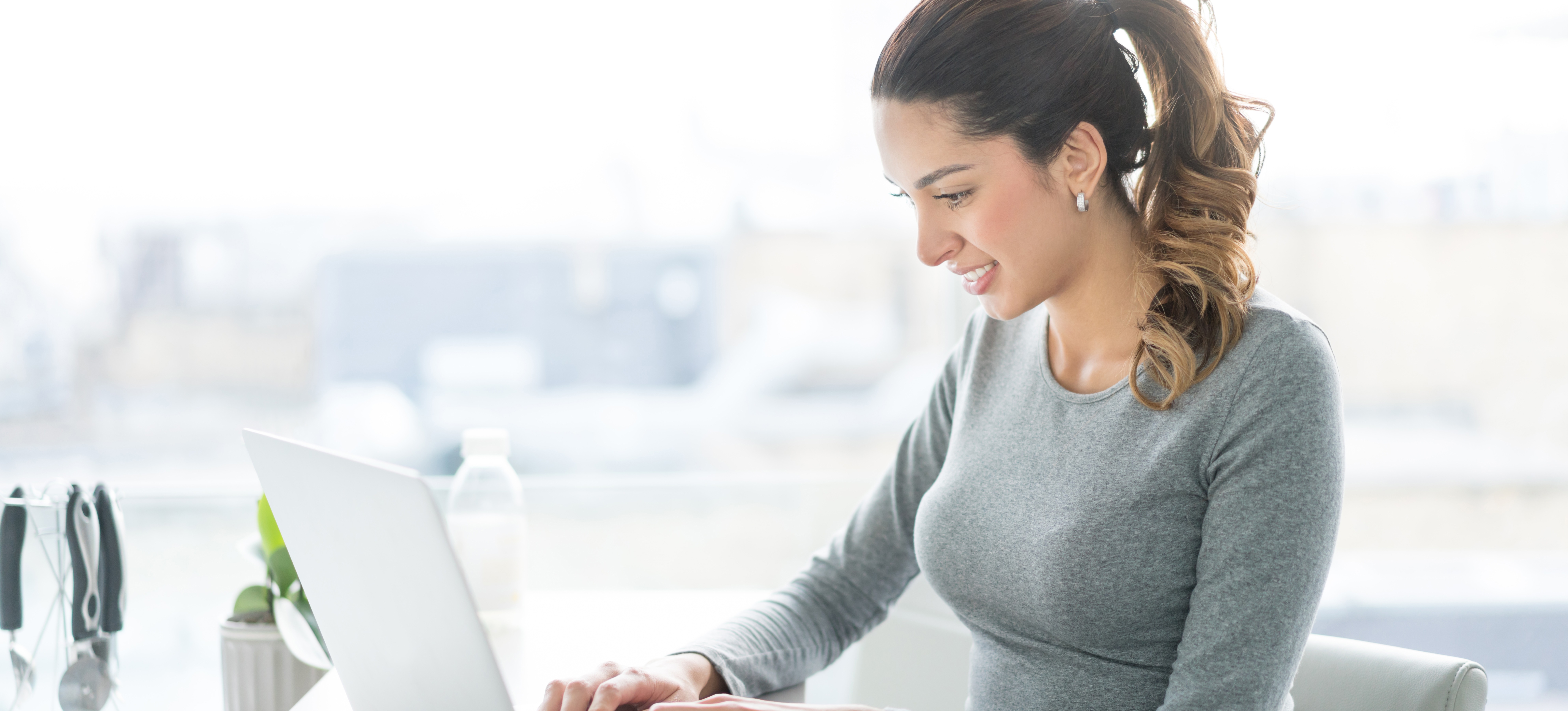 [Featured Image] A smiling woman sits at her laptop, writing an email to decline a job offer.