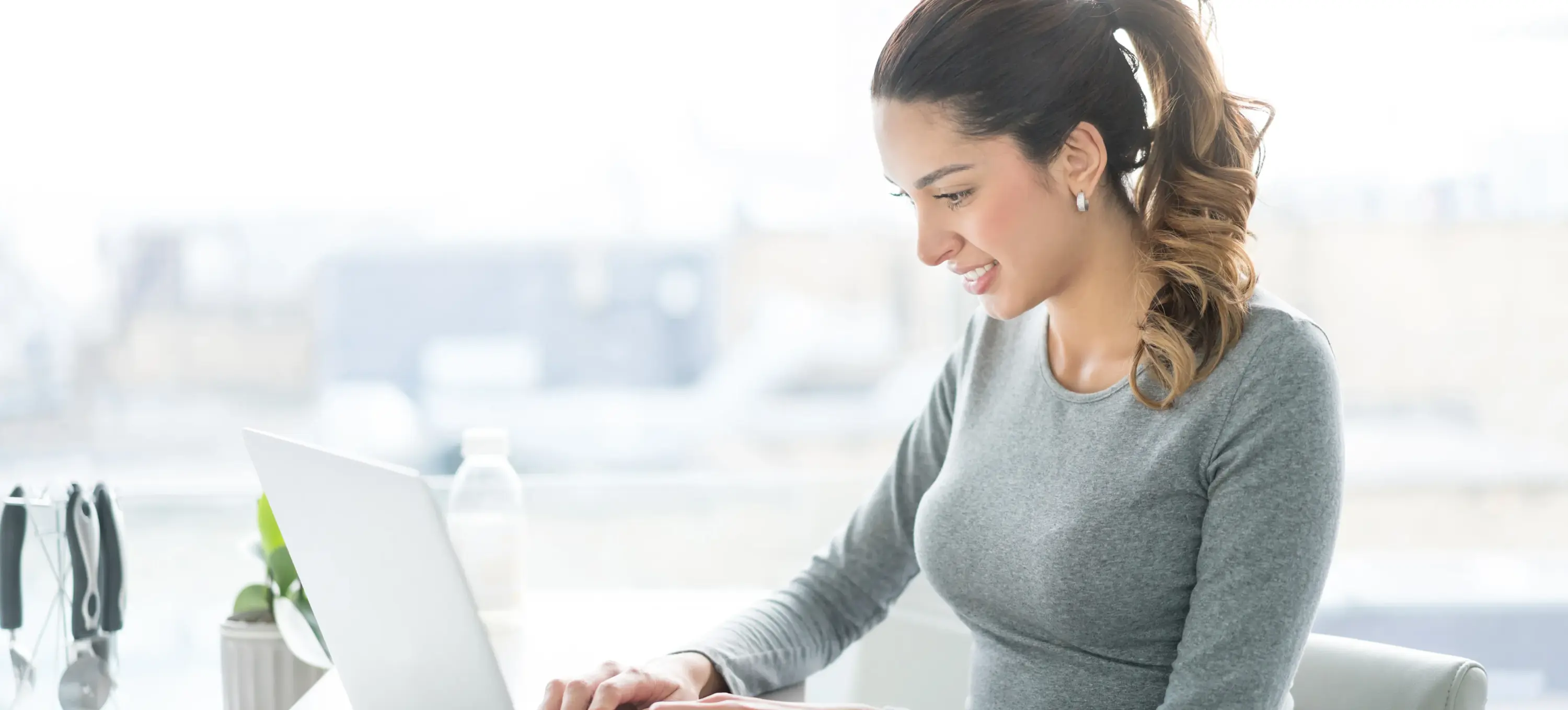 [Featured Image] A smiling woman sits at her laptop, writing an email to decline a job offer.