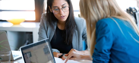 [Featured image] Two women sit at a table going over a competitive analysis report. Both of them have laptops in front of them.
