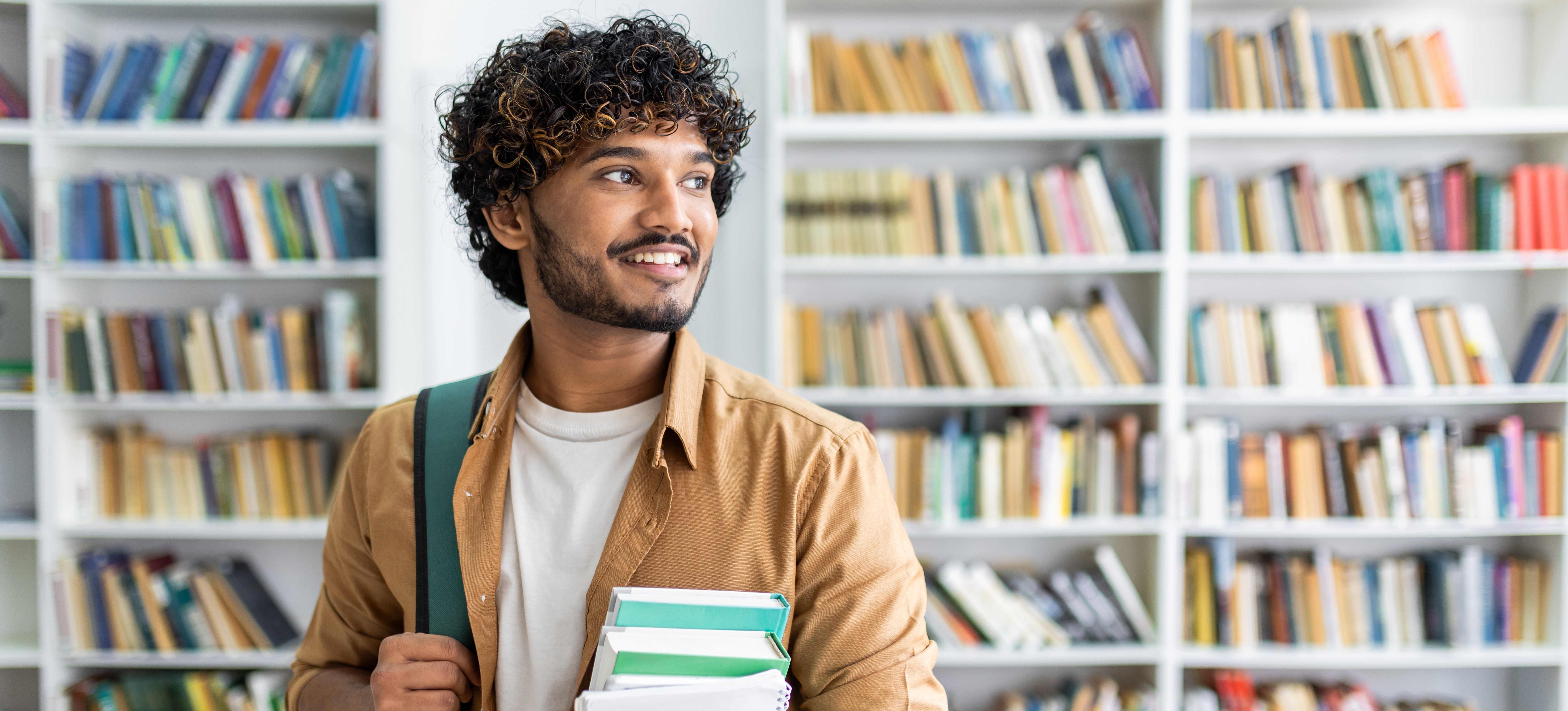[Featured Image] A student stands in a library holding books and a backpack in his Russell Group university.   