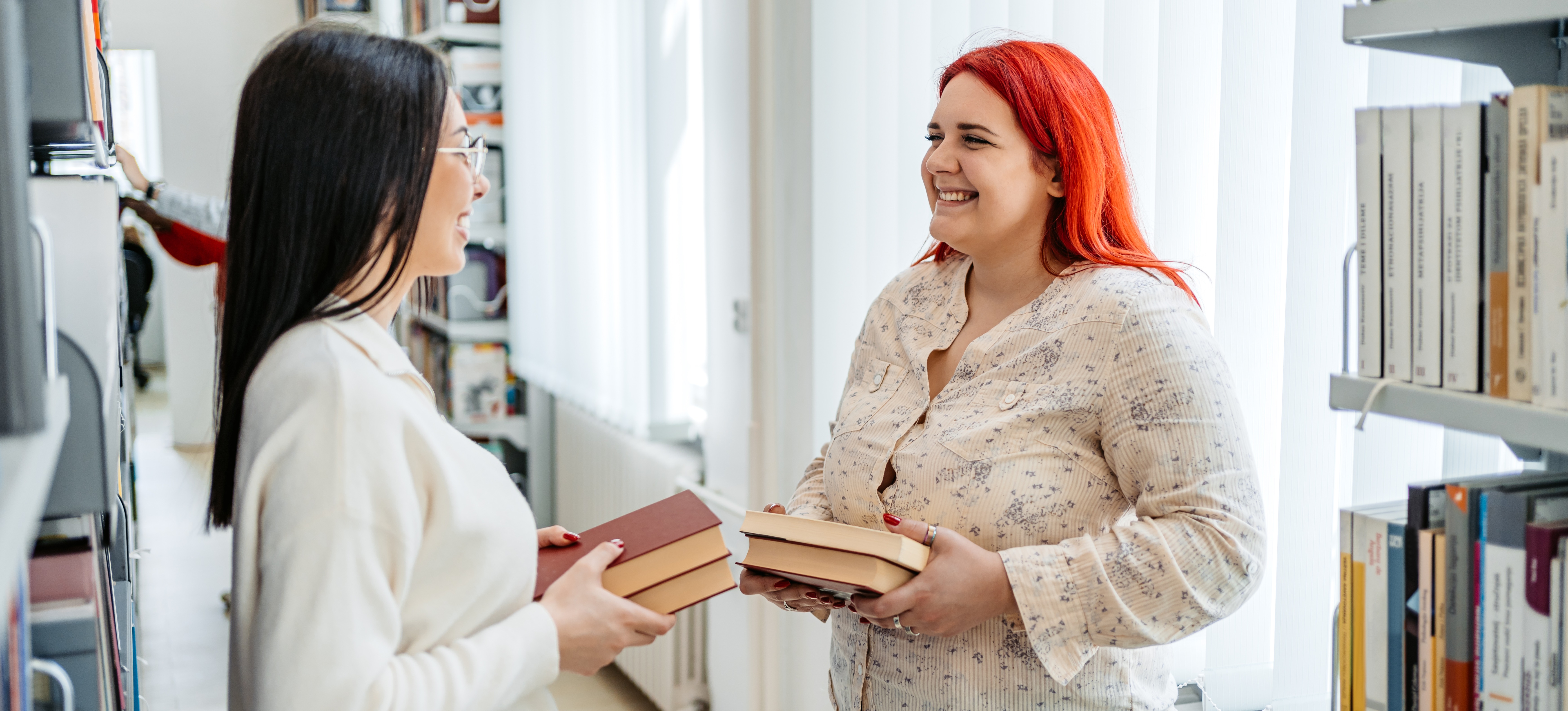 [Featured Image] Two students holding books meet in a college library to talk about an assignment while earning their MA in communication. 