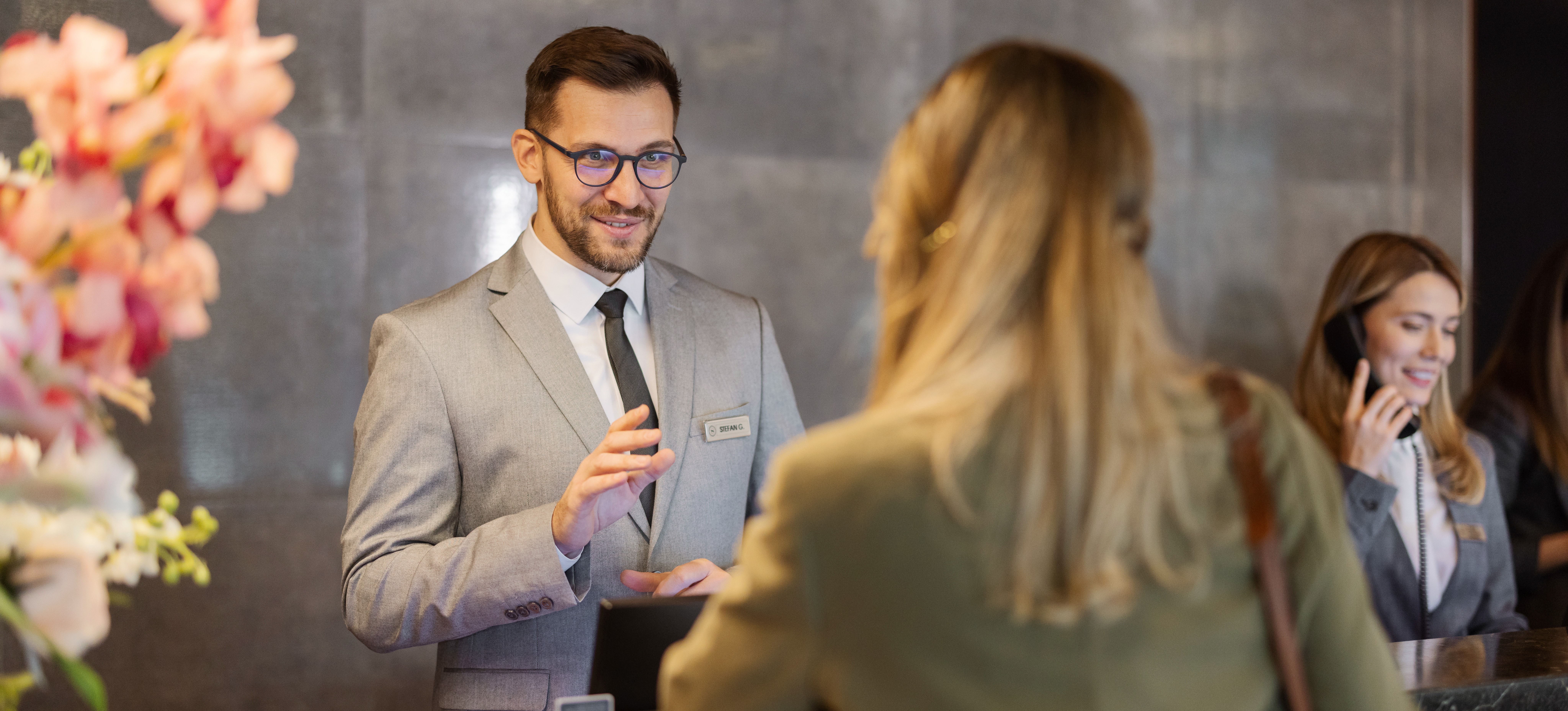 [Featured Image] A concierge who works in the hospitality industry greets a guest at the reception desk while a hotel manager talks to a potential customer on the phone.
