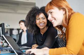 [Featured image] Two women working in IT talking to each other in the workplace.
