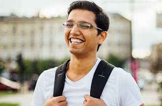 [Featured image] A young Southeast Asian man in glasses and a light blue shirt stands smiling and gripping the straps of his backpack. 