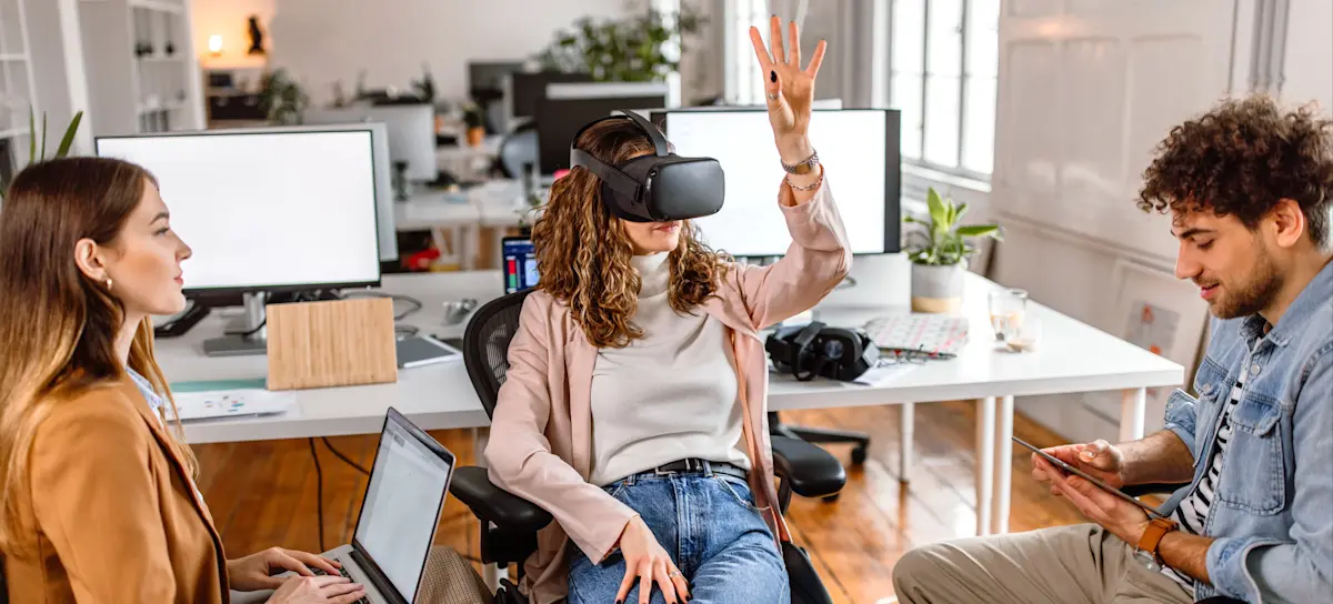 [Featured Image] A VR developer sits in an office and tests software with two colleagues.
