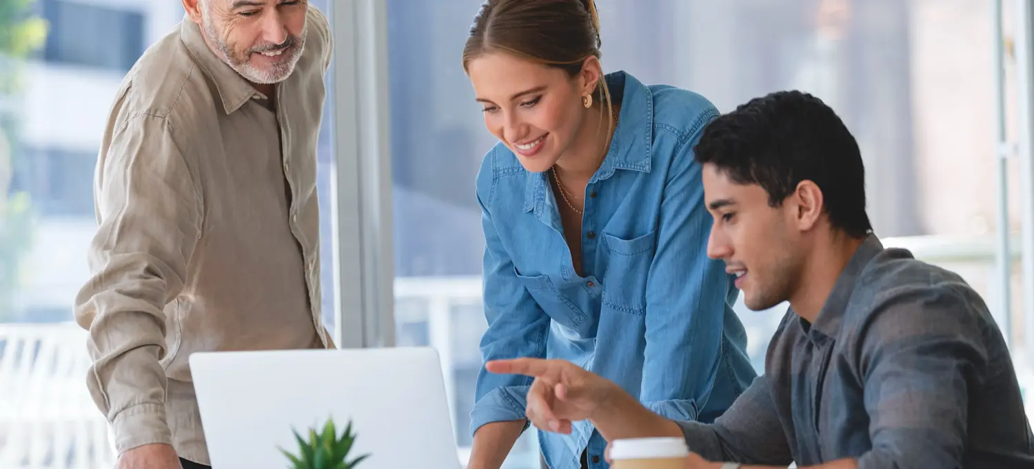 [Featured Image] Three smiling colleagues stand in an office at a desk discussing the advantages of Anthropic versus OpenAI.
