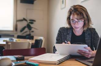 [Featured image] A young white woman with glasses stares at a tablet, while her laptop and other paper notebooks are scattered around her.