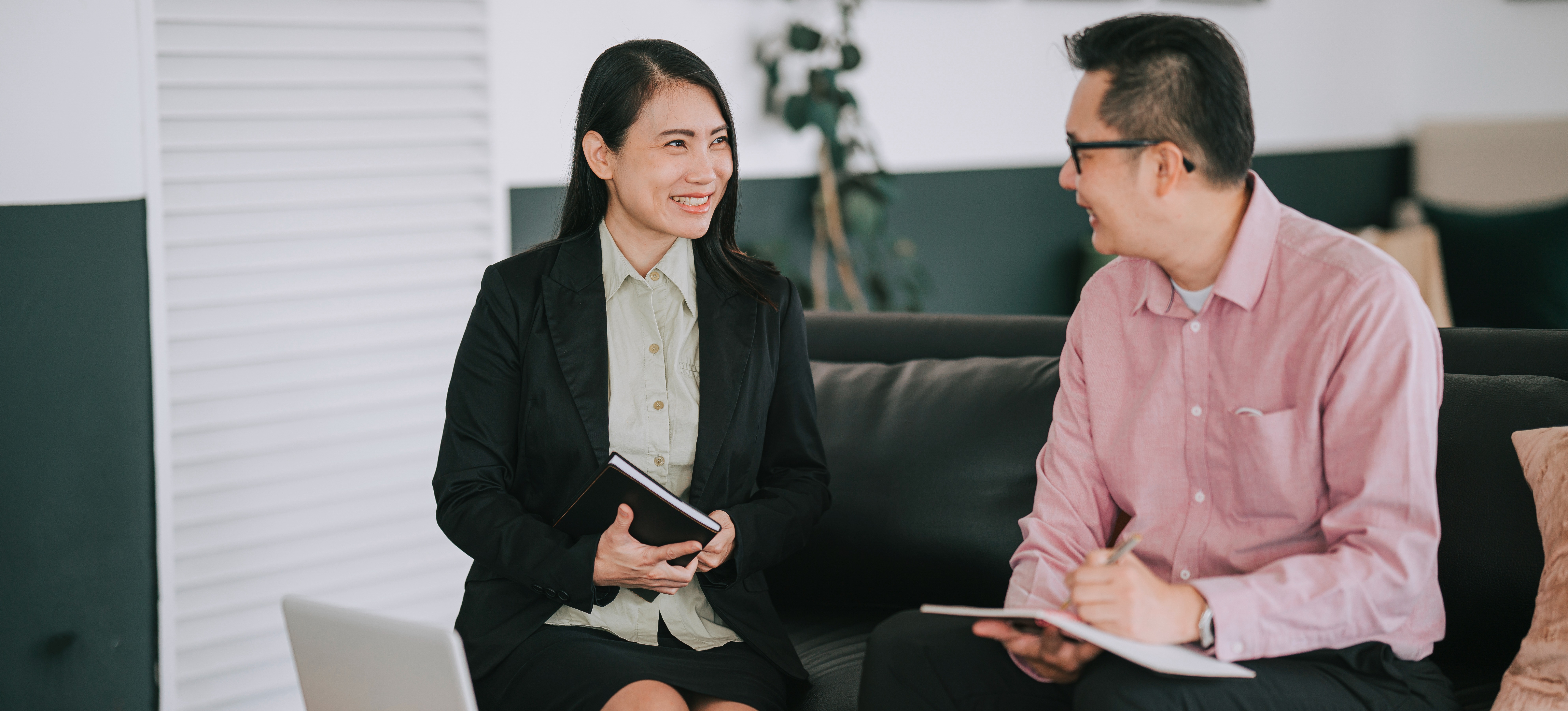 [Featured Image] A systems auditor having a discussion with a colleague in a professional workspace while seated on a sofa.