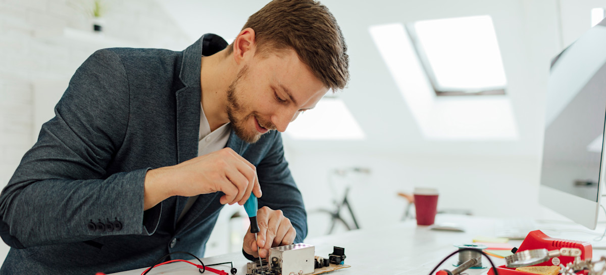 [Featured Image] In their office, a hardware engineer hovers over their desk, working on a circuit board with a screwdriver.
