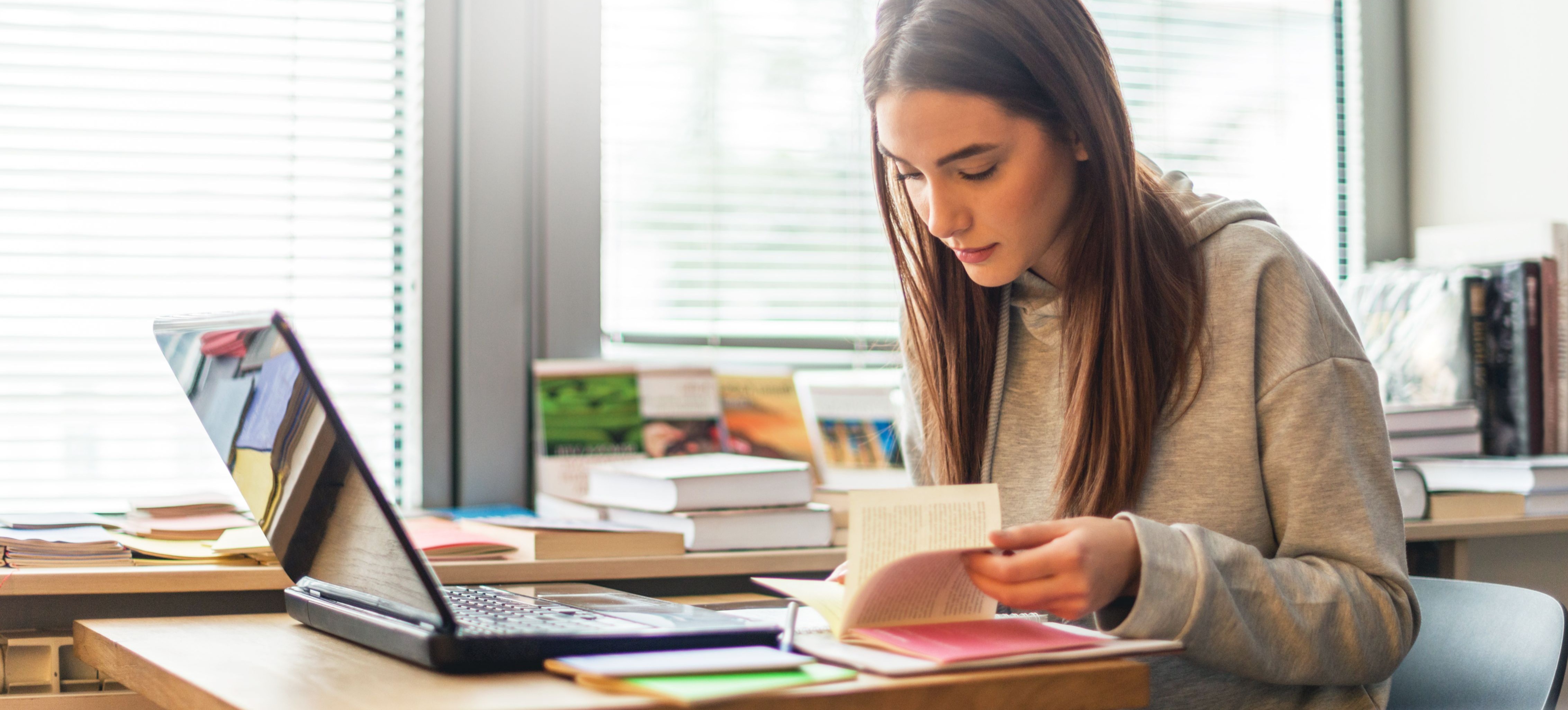 [Featured Image] A person is sitting at their desk looking through a book with a laptop in front of them.