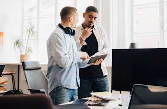 [Feautured image] Two IT support technicians discuss IT issues in front of a large monitor.