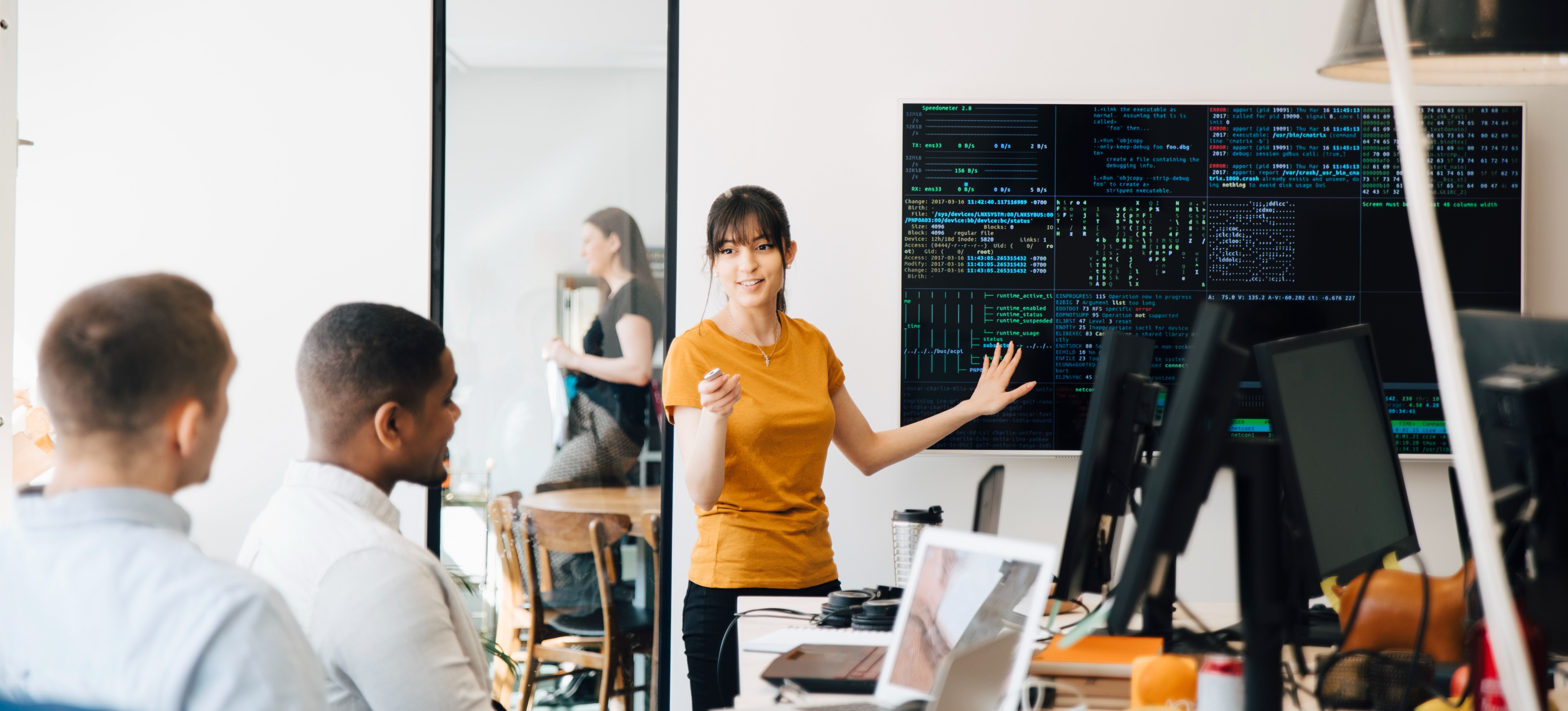 [Featured Image] A woman who followed a software developer career path presents to two men in an office with a screen filled with code behind her.
