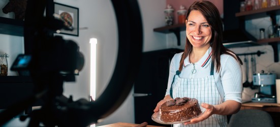 [Imagen destacada] Una influencer gastronómica sostiene un pastel y sonríe ante una cámara de vídeo.