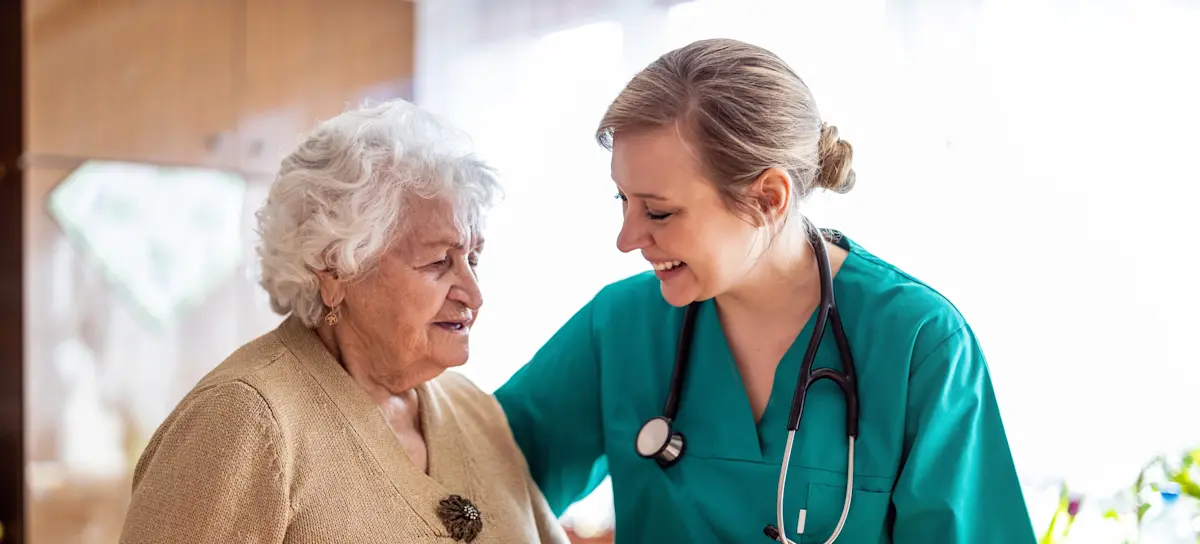 [Featured Image]: A psychiatric nurse wearing a green uniform and a stethoscope around their neck is taking care of a patient with short white hair and wearing a brown blouse. 