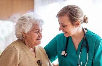 [Featured Image]: A psychiatric nurse wearing a green uniform, and a stethoscope around her neck is taking care of a patient with short white hair and wearing a brown blouse. 