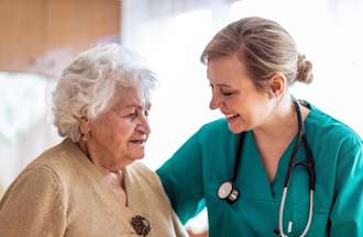 [Featured Image]: A psychiatric nurse wearing a green uniform, and a stethoscope around her neck is taking care of a patient with short white hair and wearing a brown blouse. 