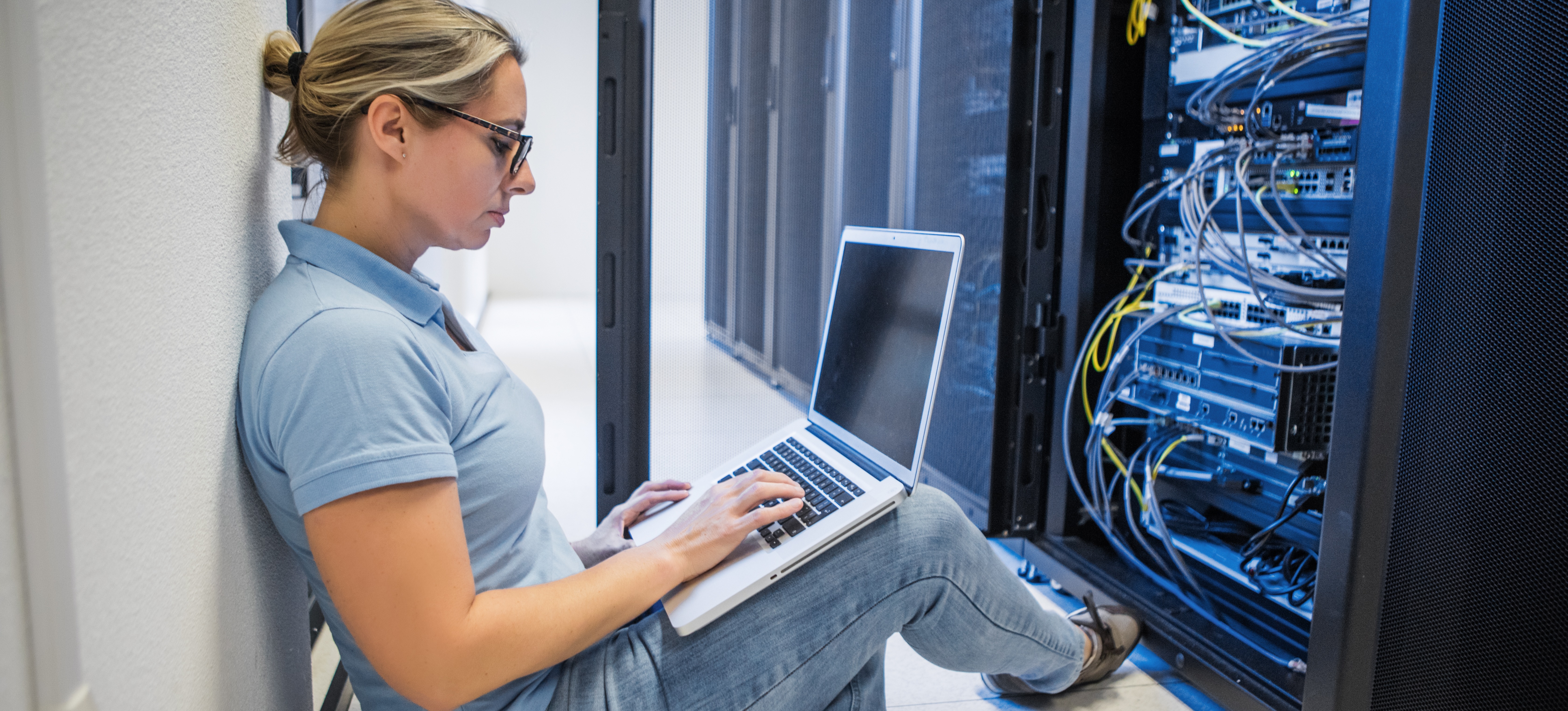 [Featured Image] A network engineer sits on a server room floor and works on their laptop as they troubleshoot network equipment in front of them.
