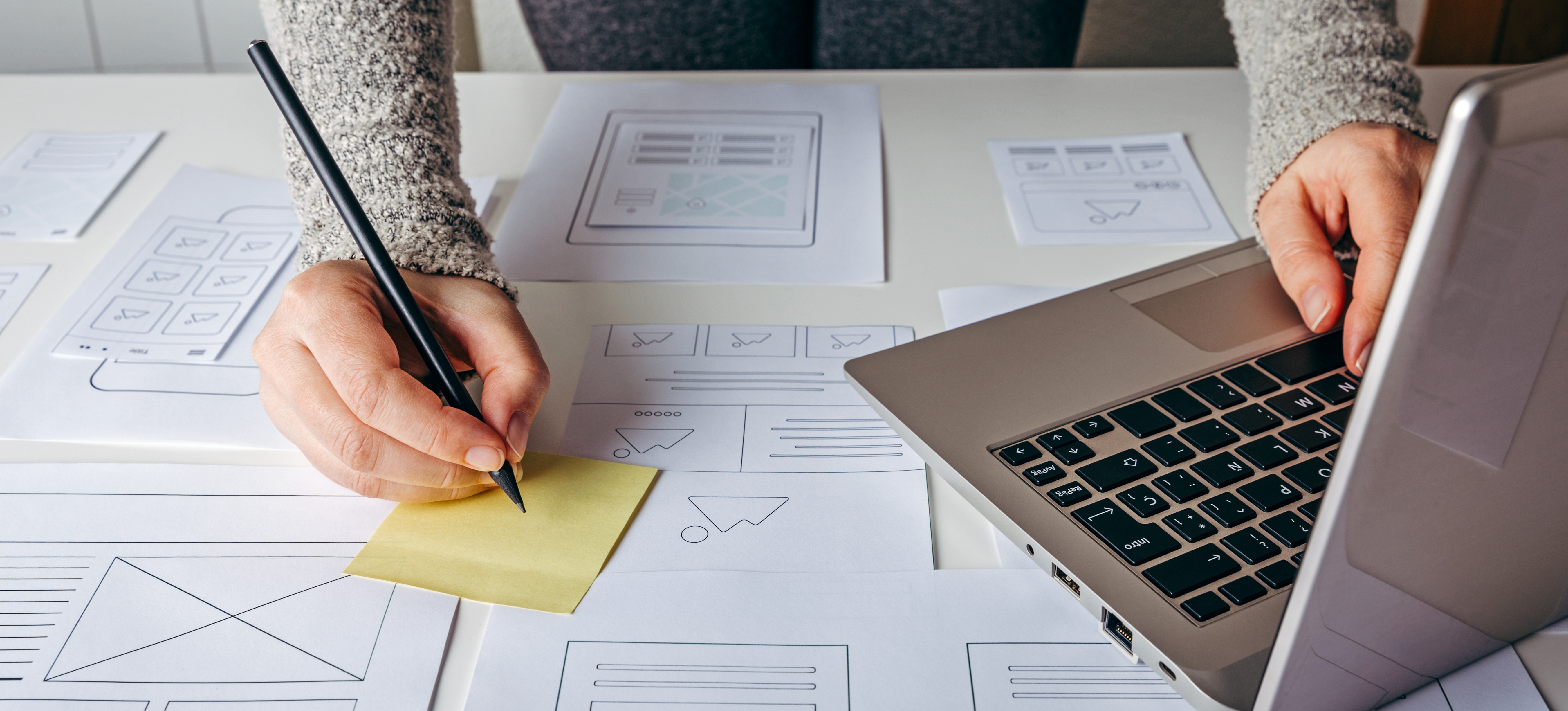 [Hauptbild] Ein UX-Designer verwendet einen Laptop und macht sich Notizen auf einem gelben Post-it-Zettel, während er Ausdrucke von Wireframes auf einem Schreibtisch überprüft.