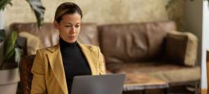 Woman wearing a blazer and turtleneck sitting in a quiet, comfortable office writing on her laptop.