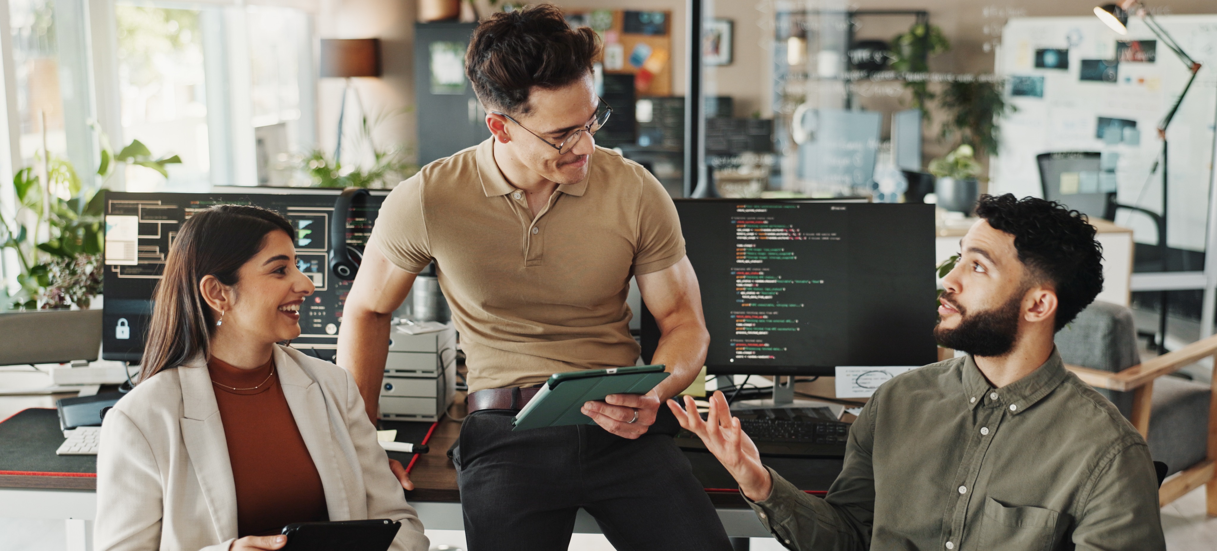 [Featured Image] Three DevOps professionals talk about infrastructure as code in an office with computer code on monitors in the background.
