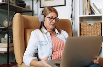 [Featured Image] A person sits in an armchair and chats using a laptop and headphones.