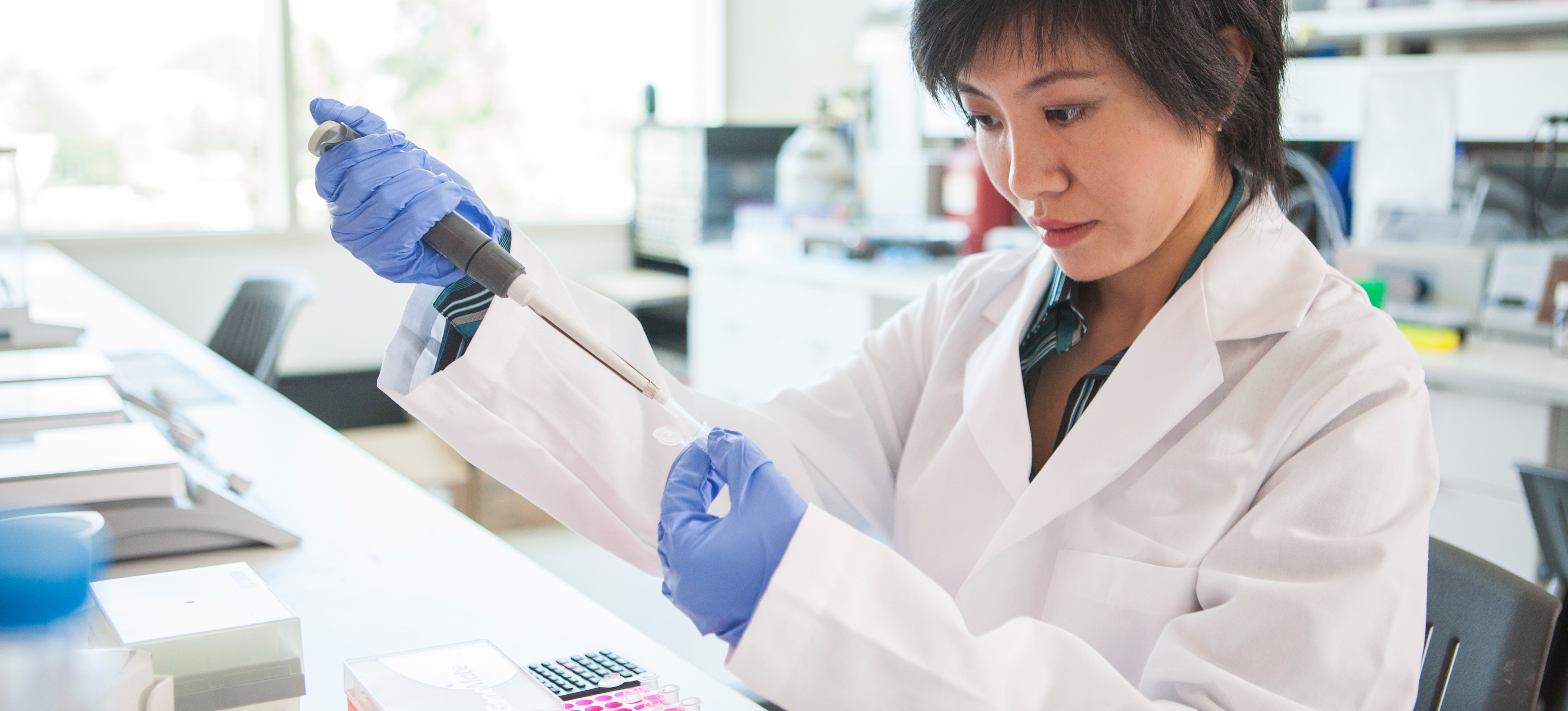 [Featured Image] An epidemiologist working in a public health career uses a syringe to add fluid to a glass slide as she studies disease control in a lab.