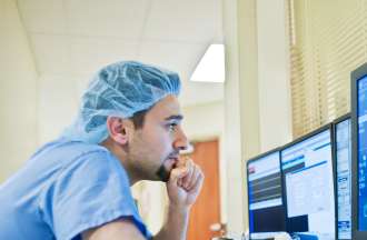 [Featured Image]: A man in blue scrubs, working in a hospital, analyzes information on his computer.