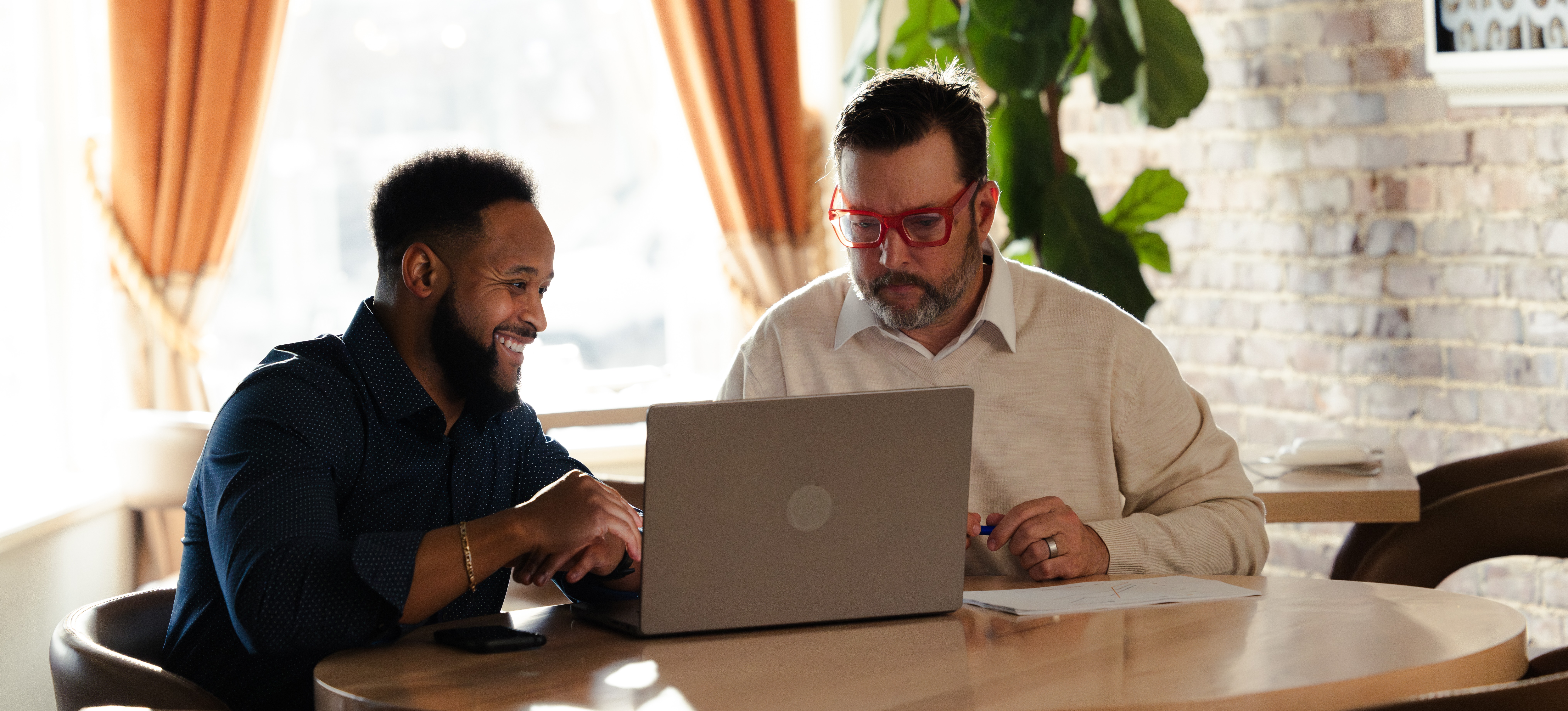 [Featured Image] Two professionals look at a laptop in a brick-walled room as they research collaborative robots and the potential for including them in their workflow.
