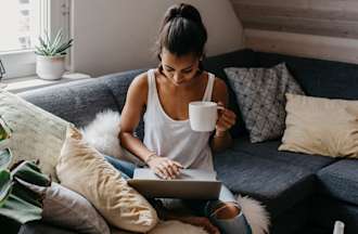 [Featured image] A woman sits on a couch, holding a white mug, as she types a data analyst cover letter on her laptop.