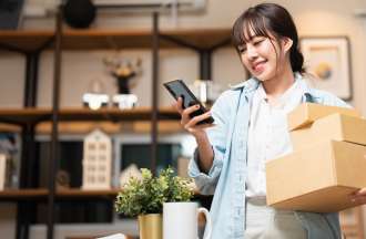 [Featured image] A woman holds three brown packages in her arm while looking at her phone. She's standing in a living room.
