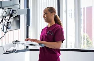 [Featured image] A nursing informatics specialist in maroon scrubs accesses a patient database at a standing workstation in a hospital.