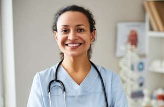 [Featured Image]:  An occupational therapy assistant, wearing a blue uniform and a stethoscope around her neck.  She is standing in an examination room, with white shelves and a white chair and desk.