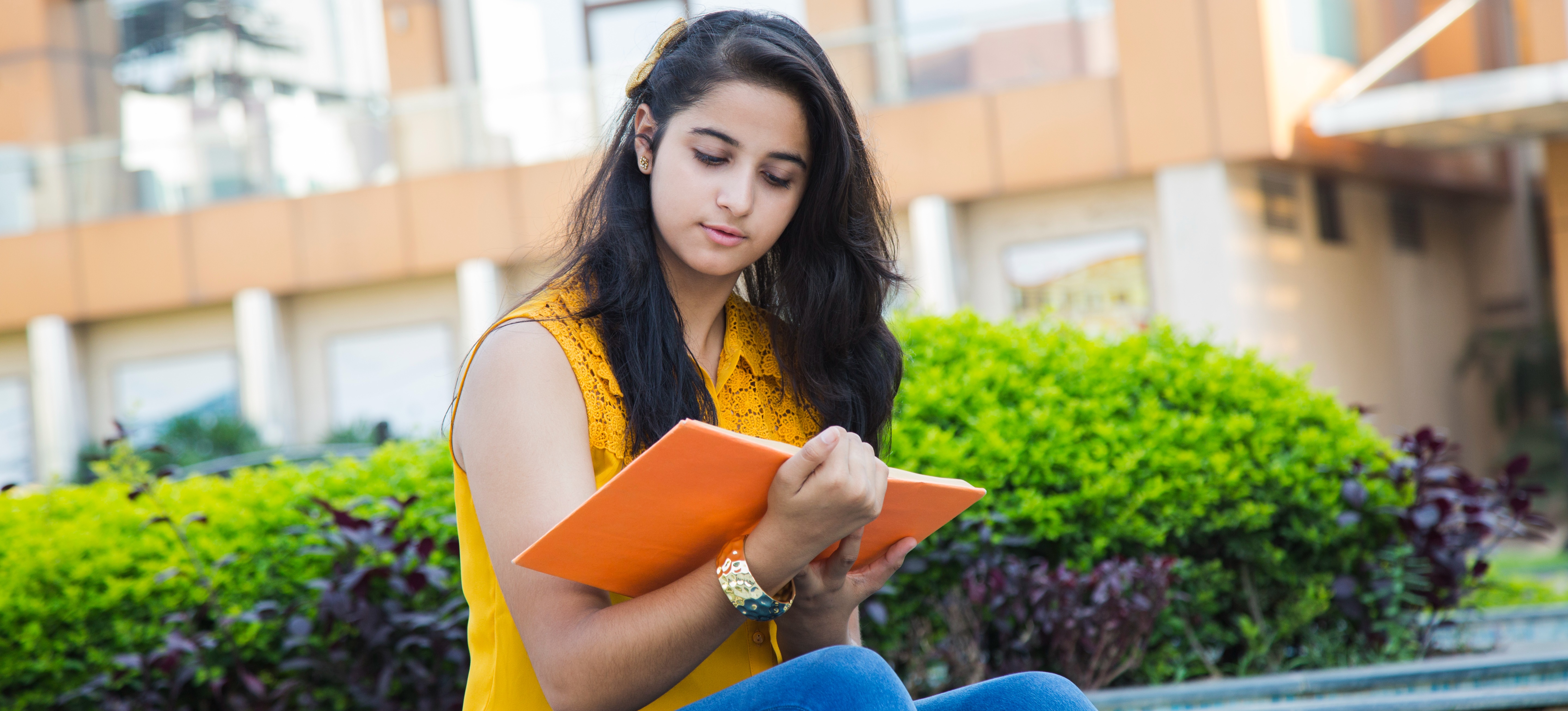 [Featured Image] A student reads a book outside her university where she is earning a Bachelor of Computer Application.   