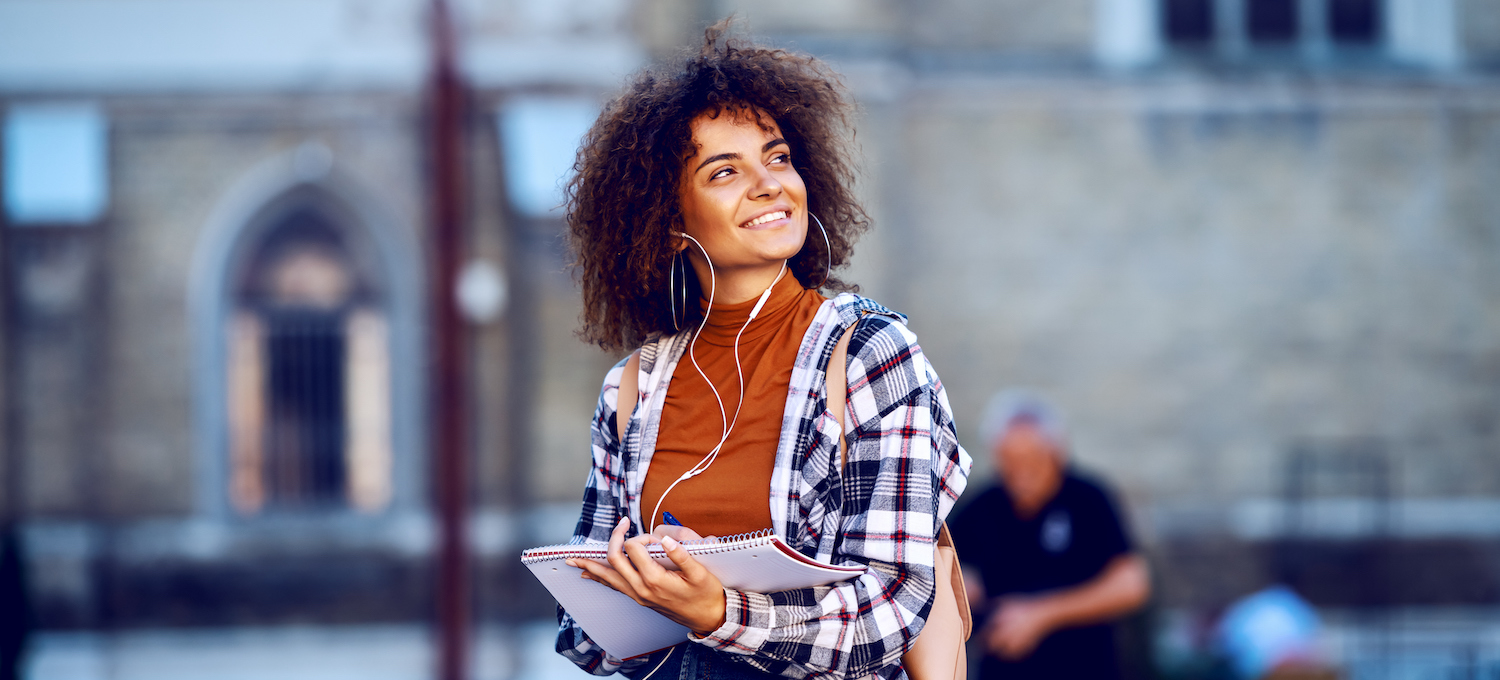 [Featured image] Student wearing a backpack and carrying a notebook in their arms looks around their college campus and smiles.