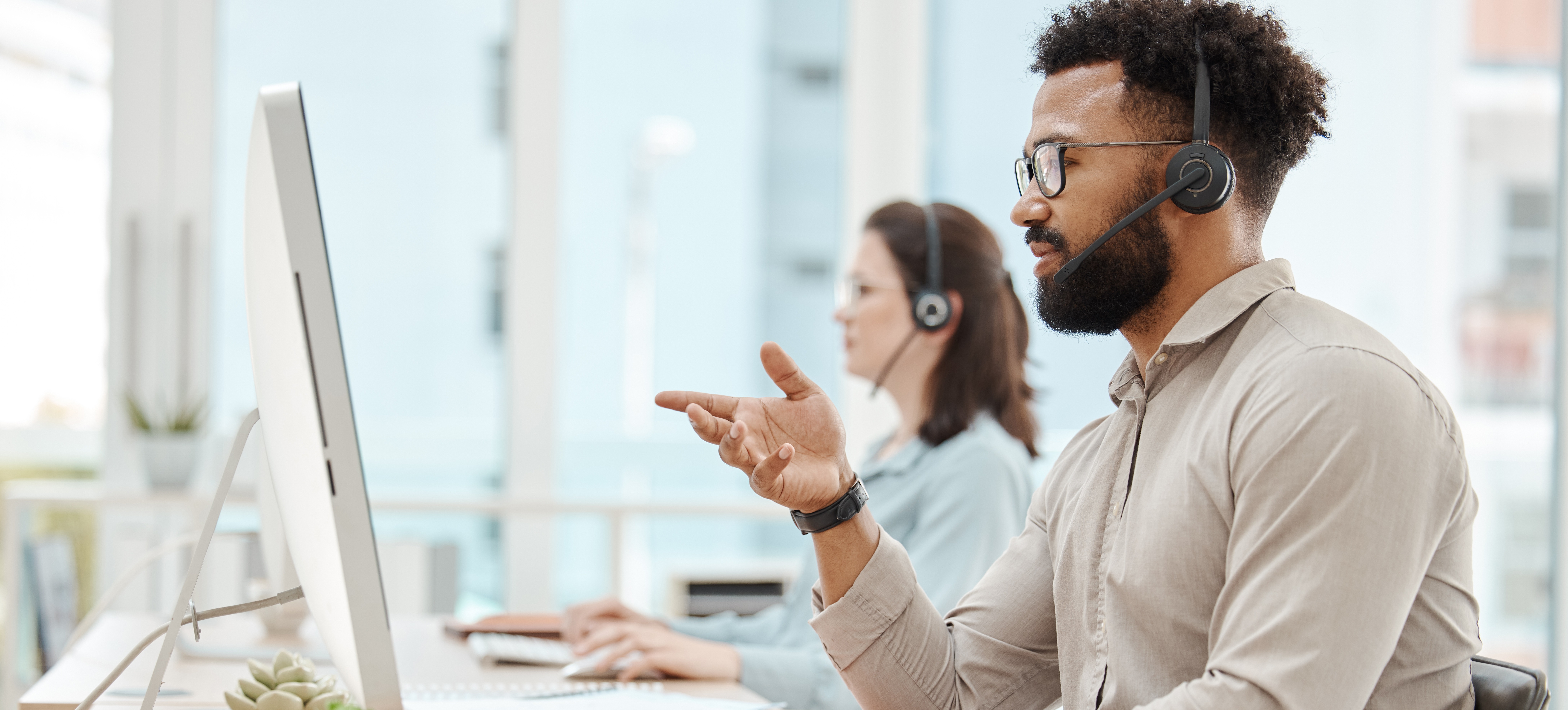 [Featured Image] A man working in IT service management talks on a headset while looking at a computer and sitting beside a colleague in a bright, airy office setting.
