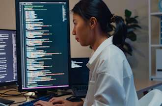 A software developer using a computer to write code sitting at a desk with multiple screens.