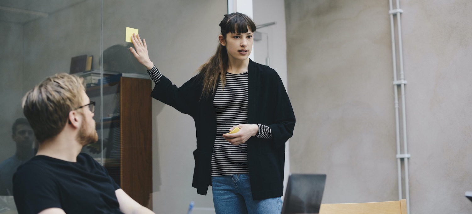 [Featured image] Two coworkers with UX certification wearing black tops work on a user design (UX) project with a laptop in a conference room with a glass wall.