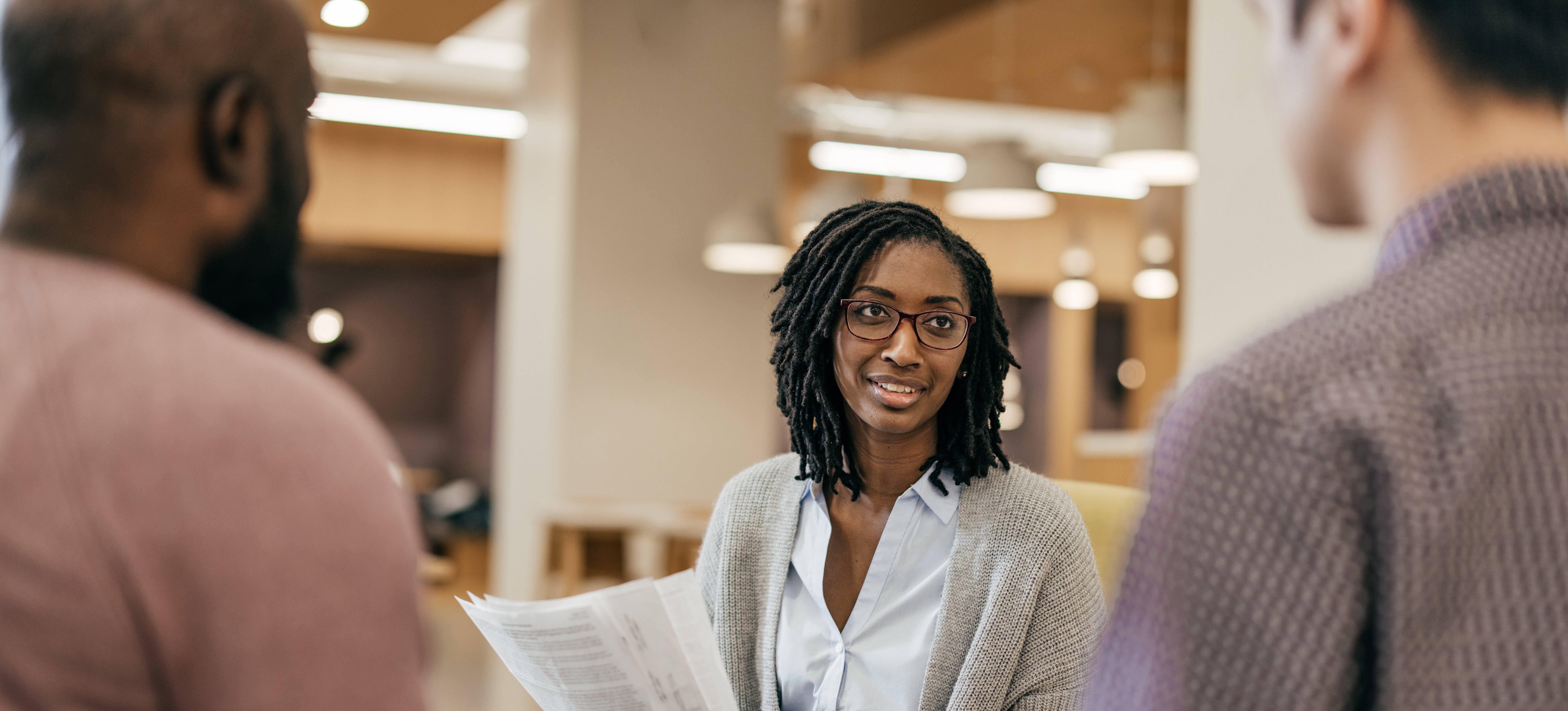 [Featured Image] A woman who learned how to become an HR manager holds a stack of papers while speaking with two colleagues in an office setting.
