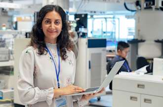 [Featured Image]:  A woman, wearing a white top and an id badge around her neck is holding and working on her laptop analyzing data in a lab.  