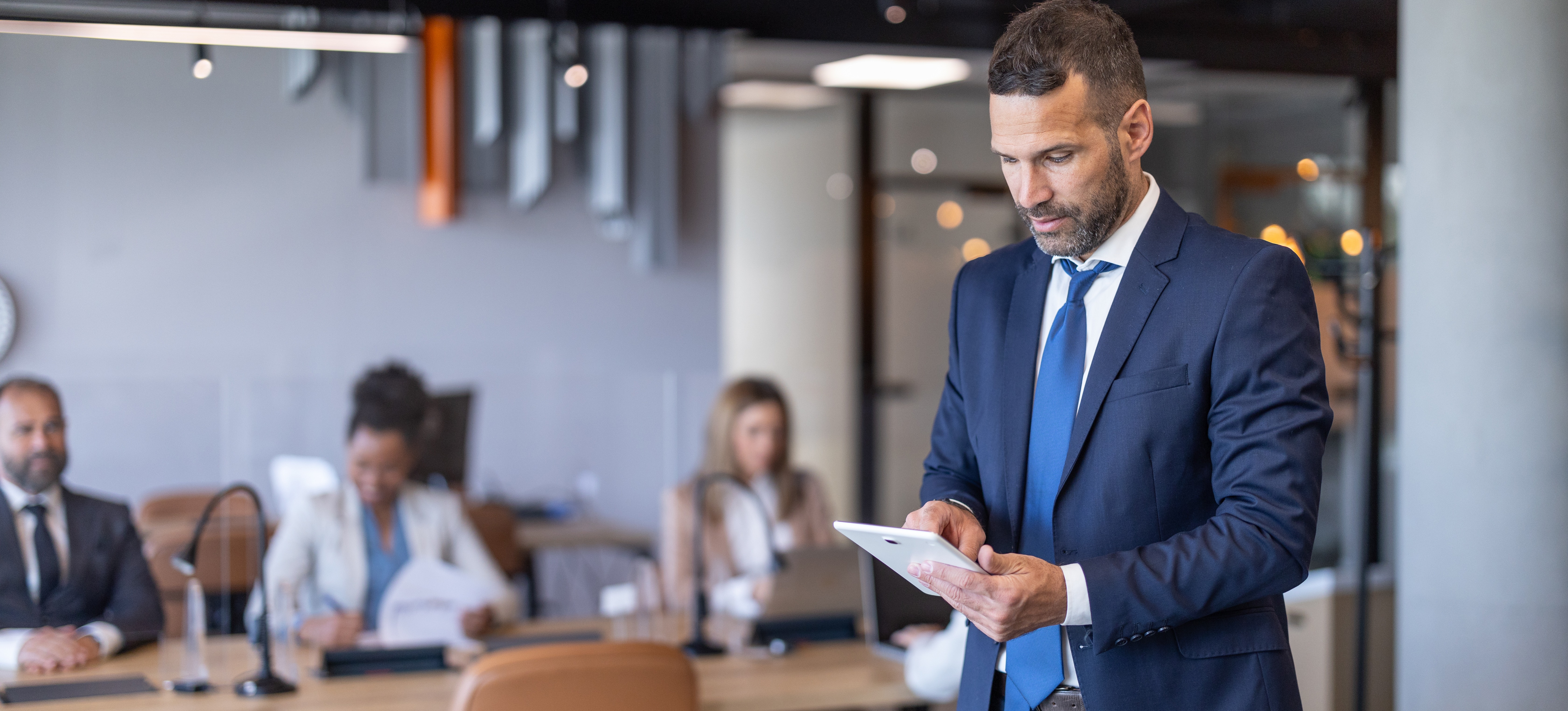 [Hauptbild] Ein Fachmann in Anzug und Krawatte steht in einem Büro und hält ein elektronisches Gerät in der Hand, während er eine Arbeitsanalyse durchführt.