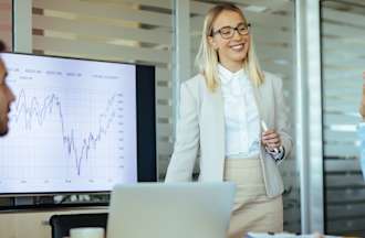 [Featured image] A marketing manager gives a presentation to company stakeholders in a conference room. She stands in front of a monitor displaying a line graph of data.