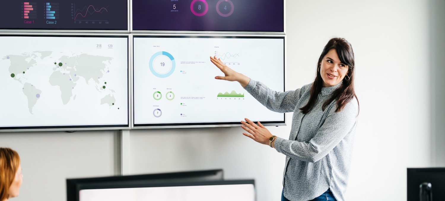 [Featured Image] A data scientist gestures to four monitors mounted on a wall that display data and presents their findings to the team. 